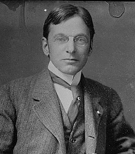 Headshot of a white man with short, brown hair; wearing a suit, tie and glasses; sitting down and looking at the camera